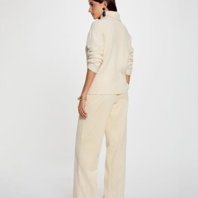 Mulher de costas com camisola creme de gola alta e calças largas creme, brincos azuis e sapatos castanhos