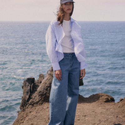 Mulher vestindo camisa azul de riscas, camisola branca, jeans largos azuis e boné azul escuro com texto, em pé perto do mar e rochas.