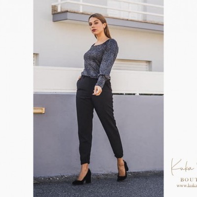 Mulher com blusa preta com padrão branco e calças pretas junto à parede cinza em ambiente urbano