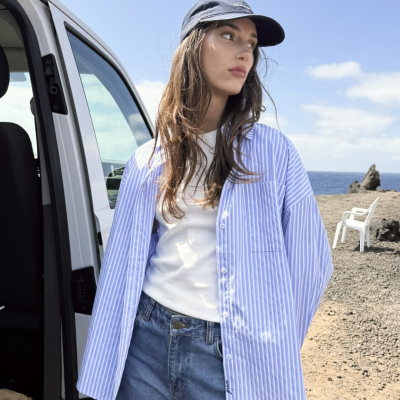 Mulher com camisa azul às riscas aberta, t-shirt branca, jeans e boné azul junto a carrinha numa paisagem costeira