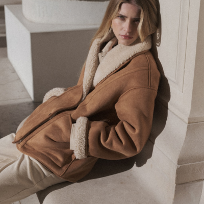 Mulher com casaco castanho de veludo com forro e gola de pelo branco creme sentada junto a uma parede