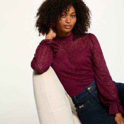 Mulher usa blusa bordô de renda e calças de ganga azul sentada numa cadeira branca