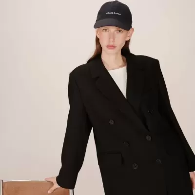 Mulher com casaco preto e boné azul escuro com texto GRACE & MILA encostada numa cadeira