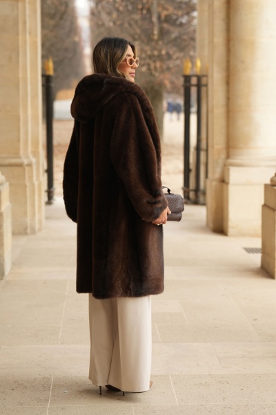 Mulher de costas com casaco de pêlo castanho escuro e calças brancas largas, segurando mala castanha pequena, em ambiente com colunas de pedra.