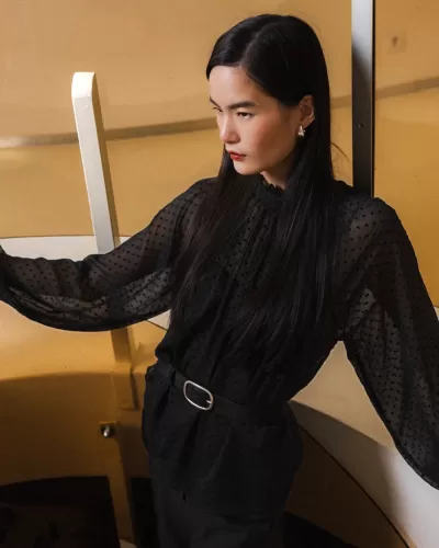 Blusa preta transparente de bolinhas com cinto preto em modelo junto a escadas interiores.