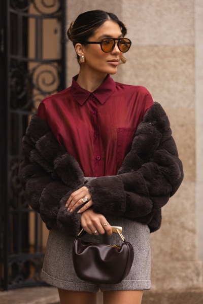 Mulher com camisa borgonha, casaco de pêlo preto, saia cinza, óculos de sol castanhos e mala preta pequena