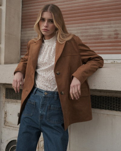 Mulher com casaco castanho, blusa branca de renda e calças de ganga azuis encostada num muro de betão