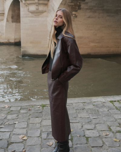Mulher com casaco e saia de couro castanho escuro junto a rio e ponte de pedra