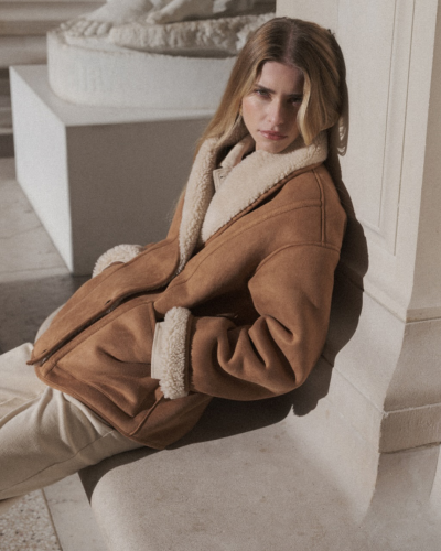 Mulher com casaco castanho de veludo com forro e gola de pelo branco creme sentada junto a uma parede