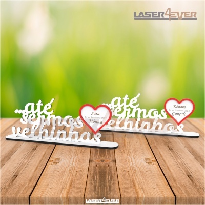 Elementos decorativos com texto 'até sermos velhinhos' e corações vermelhos com nomes, sobre superfície de madeira clara.