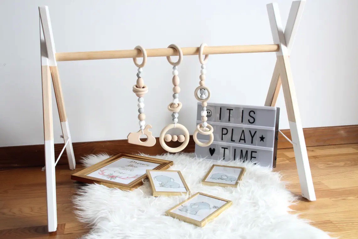 Ginásio de bebé em madeira clara e branca com brinquedos pendurados e placa de luz 'IT IS PLAY TIME'