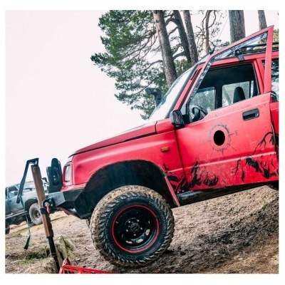 Jeep vermelho com marcas de lama e buraco na porta, pneus grandes, em ambiente natural com árvores