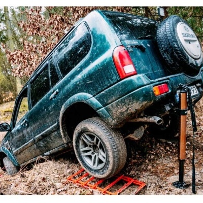 SUV verde com pneu sobresselente Suzuki em terreno acidentado com macaco hidráulico e apoios vermelhos