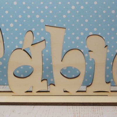 Nome em madeira 'Fábio' recortado sobre base, fundo azul com bolinhas brancas