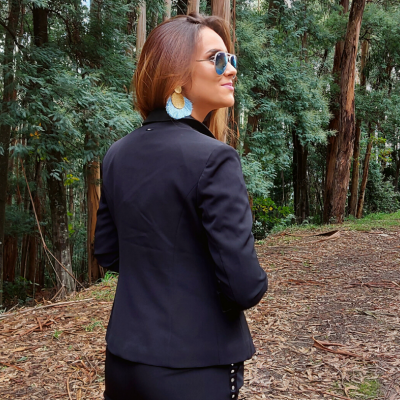 Mulher de casaco preto, brincos grandes e óculos de sol em ambiente florestal.