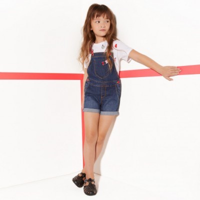 Criança vestindo macacão curto de ganga azul e t-shirt branca estampada com cerejas em fundo branco com faixa vermelha