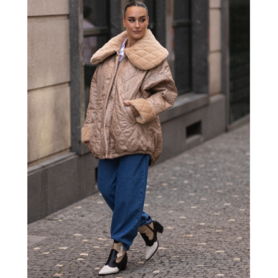 Mulher a usar casaco acolchoado bege, calças de ganga azuis largas e botins preto e branco numa rua de pedra.
