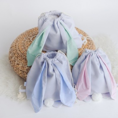 Três sacos em tecido azul claro decorados como coelhos com orelhas coloridas e pompons, sobre tapetes, em fundo claro.