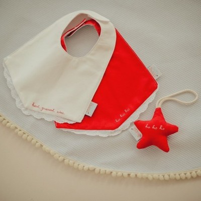 Babete branco e vermelho com brinquedo estrela vermelho sobre manta bege com pompons.