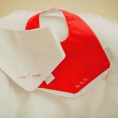 Dois babetes para bebés, um branco com bordado vermelho e outro vermelho com bordado branco sobre almofada branca