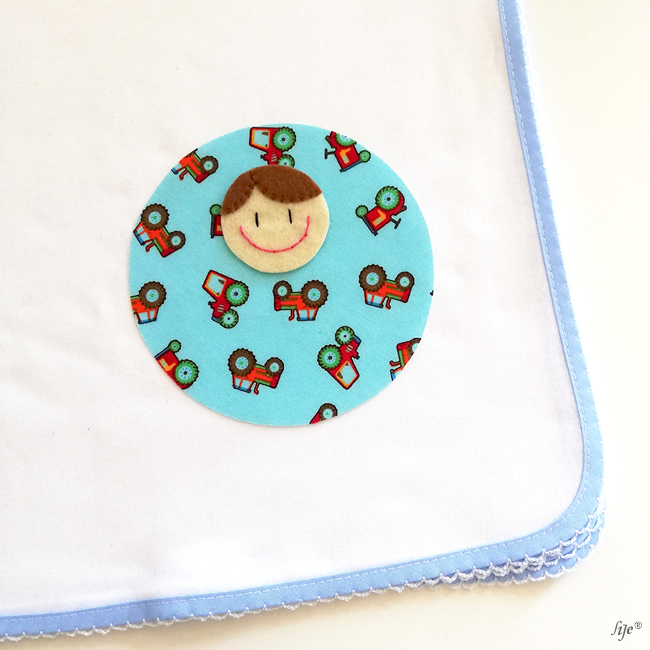 Fralda branca com borda azul e aplicação circular azul com padrão de carros e rosto de bebé em feltro.