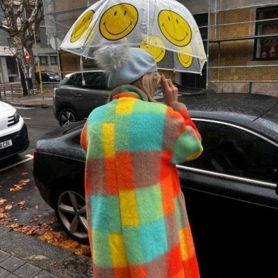 Pessoa de costas com casaco quadriculado colorido, guarda-chuva transparente com carinhas sorridentes e gorro cinza com pompom