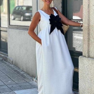 Vestido longo branco com flor preta aplicada, mulher com óculos escuros e sandálias pretas junto a edifício urbano