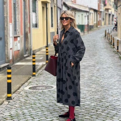 Mulher com vestido cinza com bolas pretas e acessórios vinhos numa rua de calçada de pedra