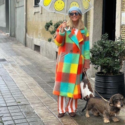 Mulher com casaco colorido e guarda-chuva transparente com carinhas felizes a passear com cão numa rua molhada