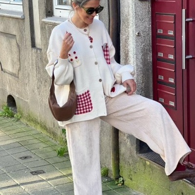 Mulher com cardigan branco de botões e bolsos xadrez, calças largas bege, bolsa castanha, cabelo apanhado com laço, óculos escuros, junto a porta vermelha e parede de pedra