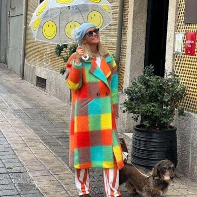 Mulher com casaco colorido e guarda-chuva transparente com carinhas sorridentes a passear na rua com cão pequeno.