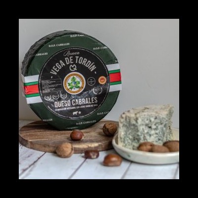 Queijo Cabrales redondo embalado com etiqueta preta e verde, fatia de queijo azul e castanhas em base de madeira.
