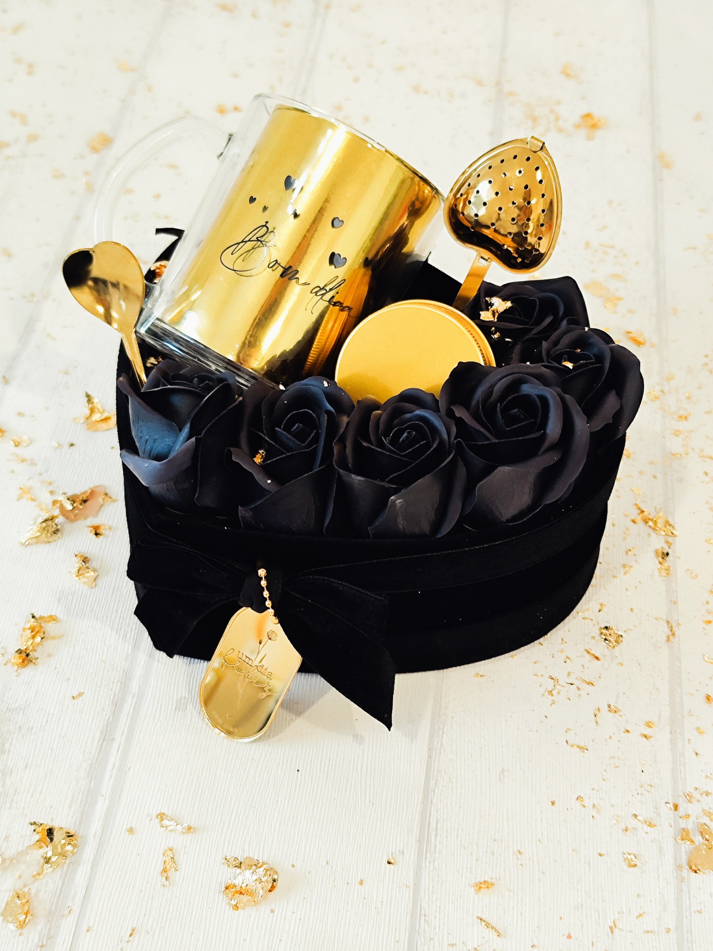 Conjunto de chá dourado com caneca, colher e infusor em caixa preta com rosas negras.