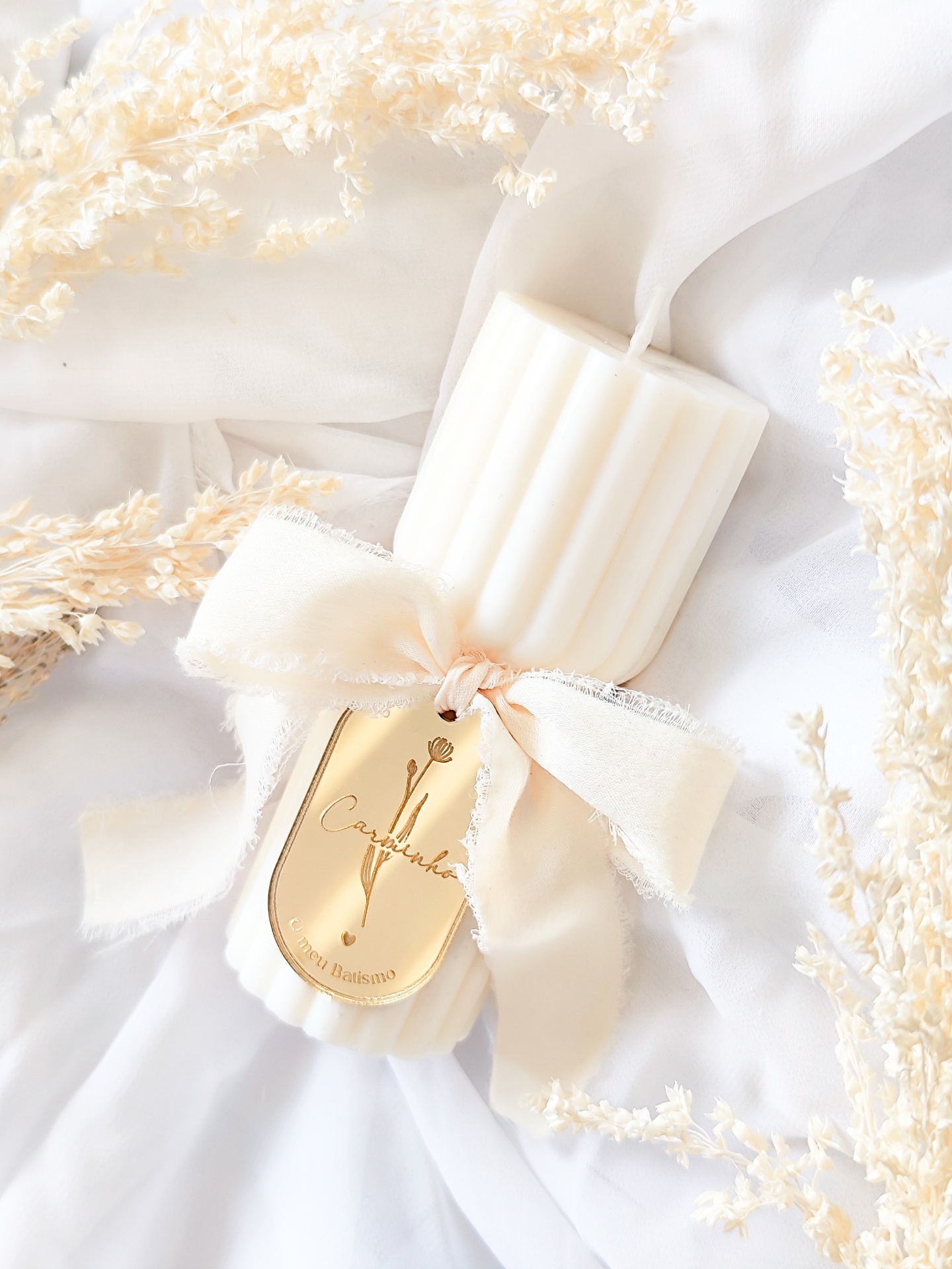 Vela branca canelada com fita creme e etiqueta dourada sobre tecido branco com flores secas