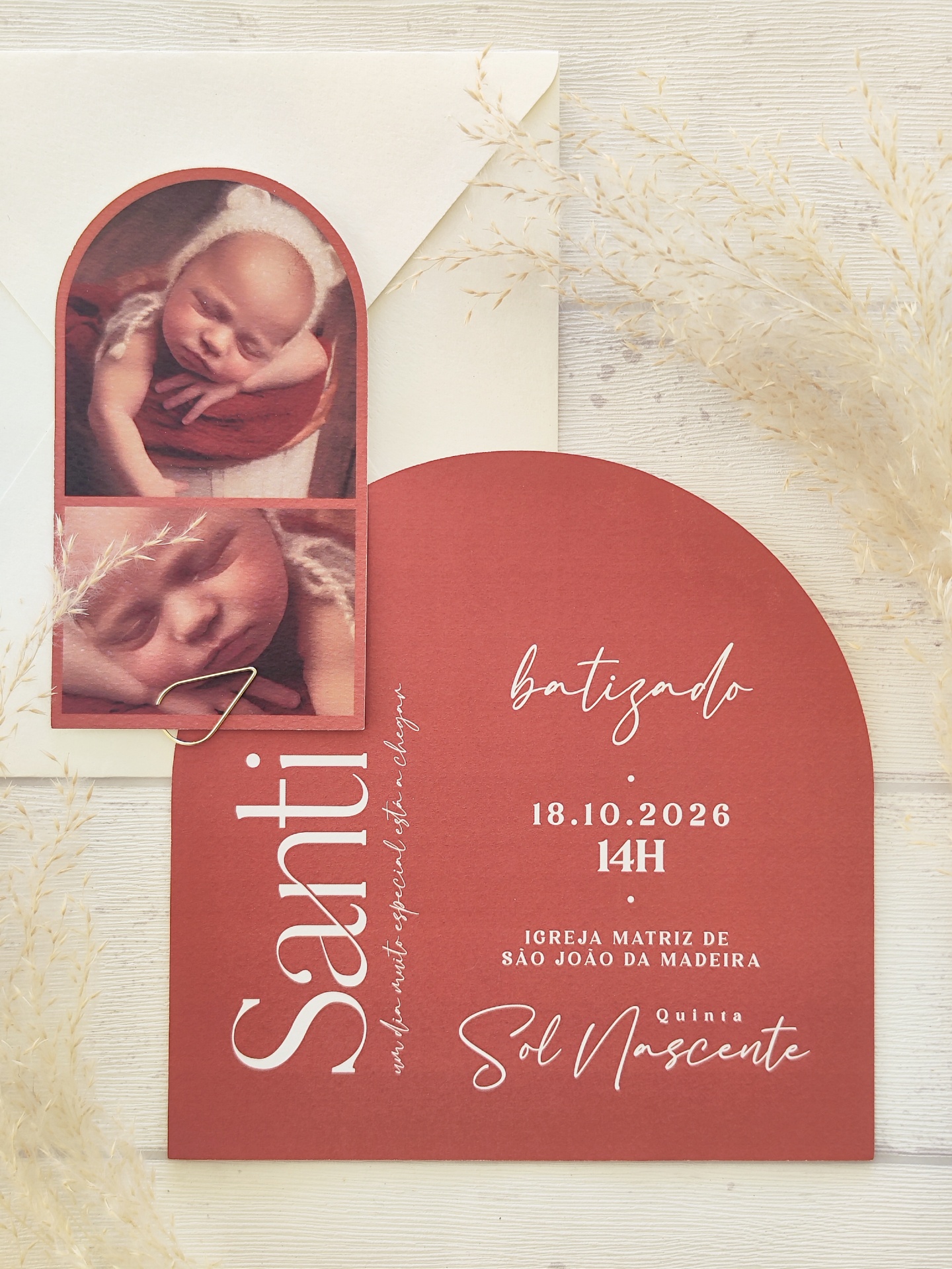 Convite batizado vermelho com fotos de bebé e envelope branco com ramos secos.