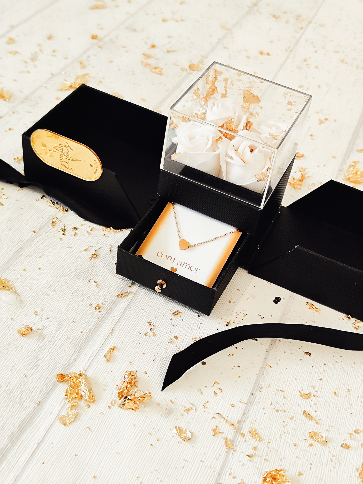 Colar com coração dourado em caixa preta com rosas brancas e papel dourado no fundo branco