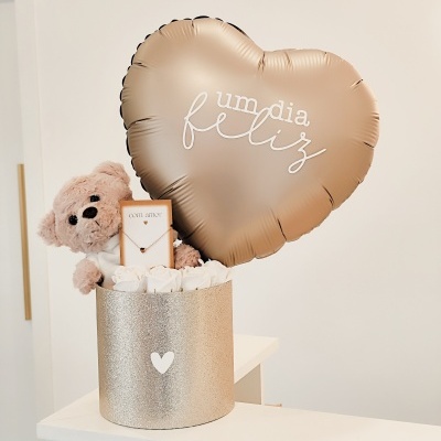 Conjunto de presente com balão dourado em forma de coração, ursinho, colar e flores brancas numa caixa dourada.