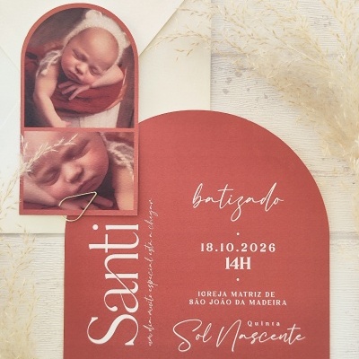 Convite batizado vermelho com fotos de bebé e envelope branco com ramos secos.