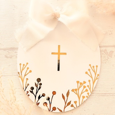 Cartão oval branco com laço e cruz dourada sobre madeira clara