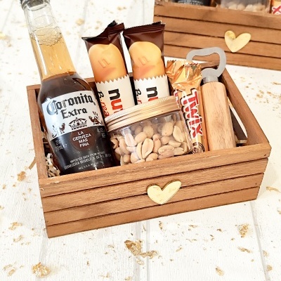 Caixa de madeira com coração dourado e produtos variados incluindo cerveja, chocolates e amendoins.