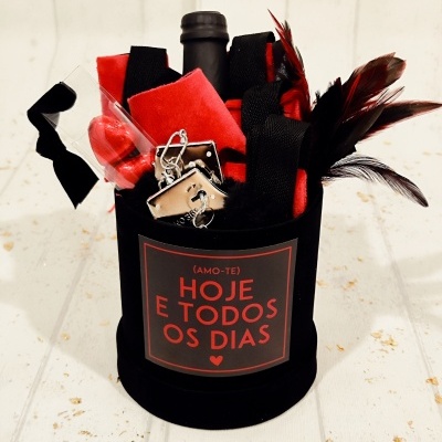 Conjunto de presentes com garrafa, penas, algemas e tecido em caixa preta com etiqueta vermelha