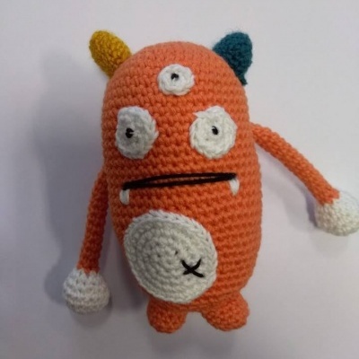 Boneco de tricot laranja com olhos e detalhes brancos, e chumaços coloridos no topo