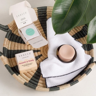 Cesto trançado com toalha, sabonete e embalagens SHAECO, com folhas verdes ao fundo.