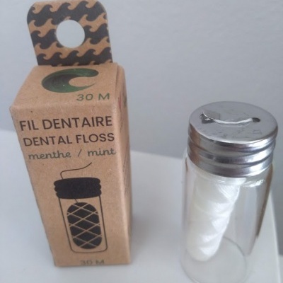 Embalagem castanha clara de fio dentário com texto FIL DENTAIRE DENTAL FLOSS e recipiente transparente com tampa prateada contendo fio dentário
