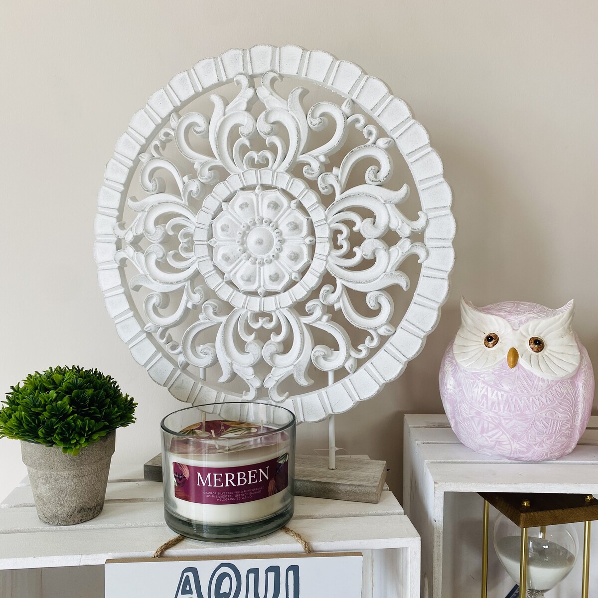 Conjunto decorativo com vaso, vela MERBEN, escultura branca detalhada e coruja rosa sobre móveis brancos.