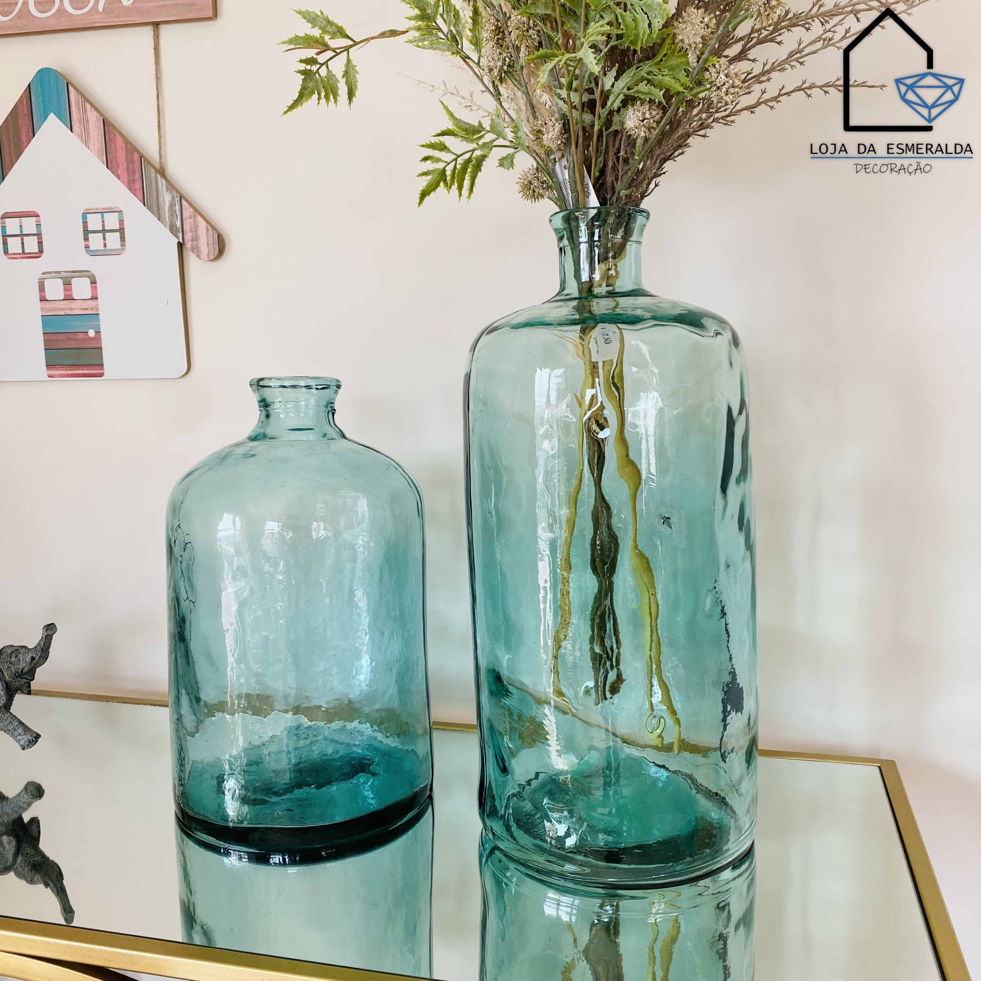 Duas garrafas decorativas de vidro verde água numa superfície espelhada, com plantas secas e decoração ao fundo