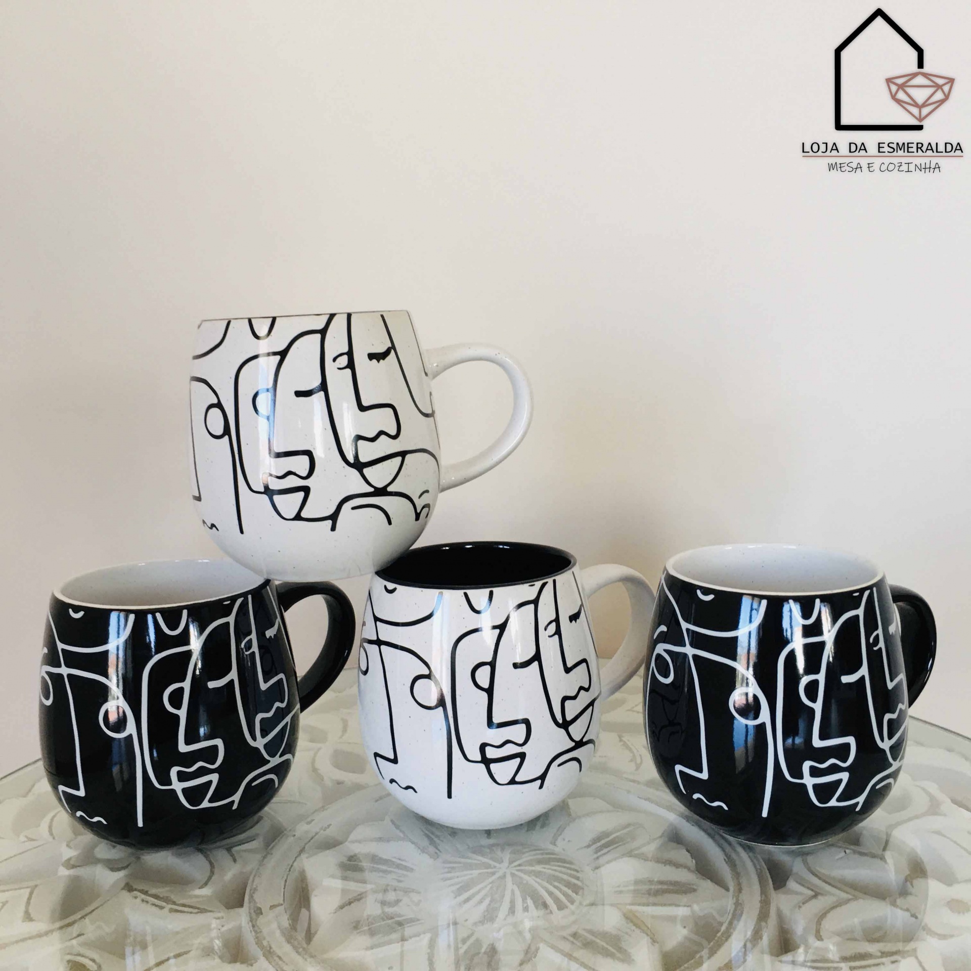 Quatro canecas de cerâmica preta e branca com padrão abstrato sobre superfície decorada e logotipo ao fundo.