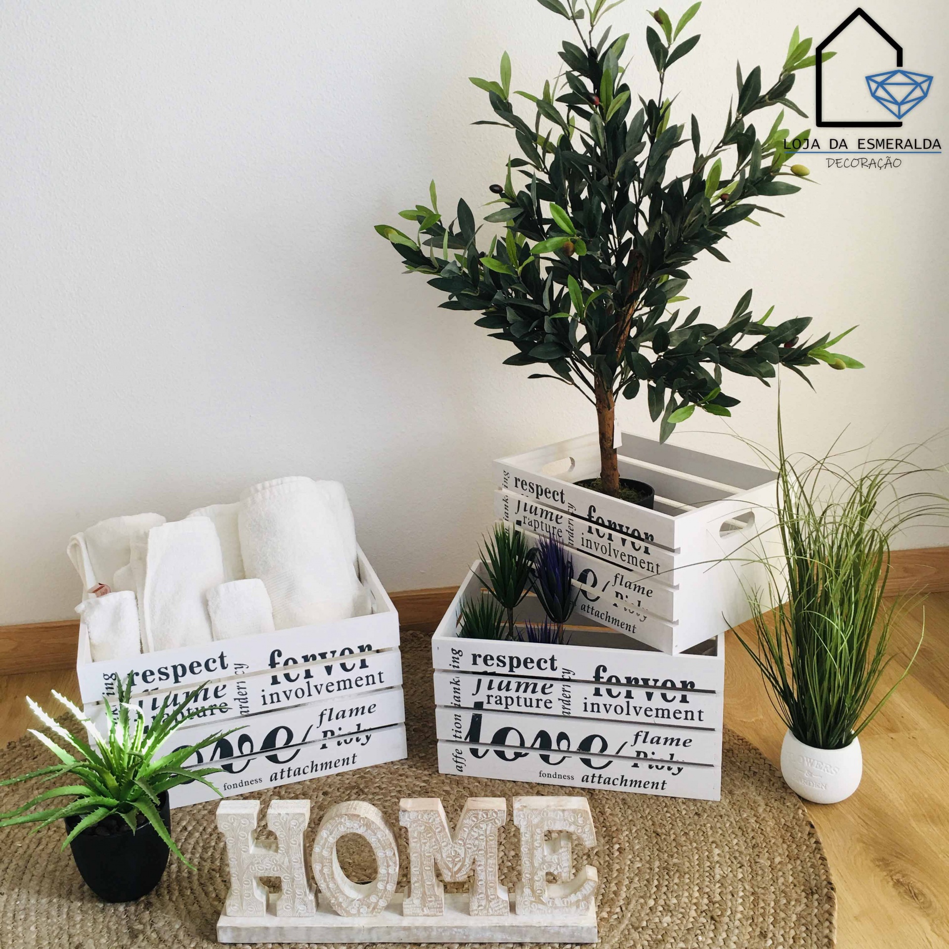 Caixas de madeira brancas decorativas com texto e plantas artificiais, toalhas e garrafas, em tapete de fibra e piso de madeira clara