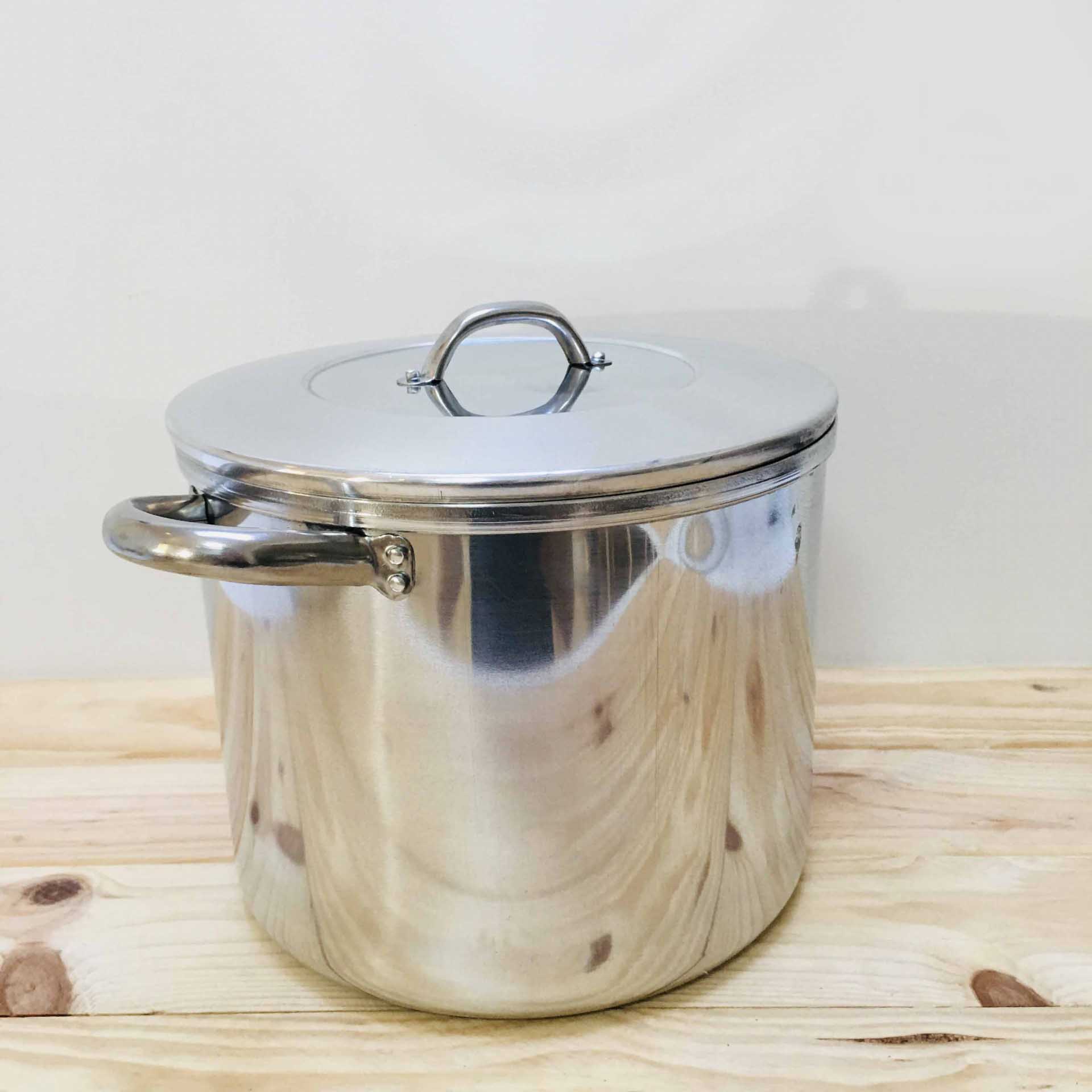 Panela em aço inoxidável com tampa e pegas em superfície de madeira clara.