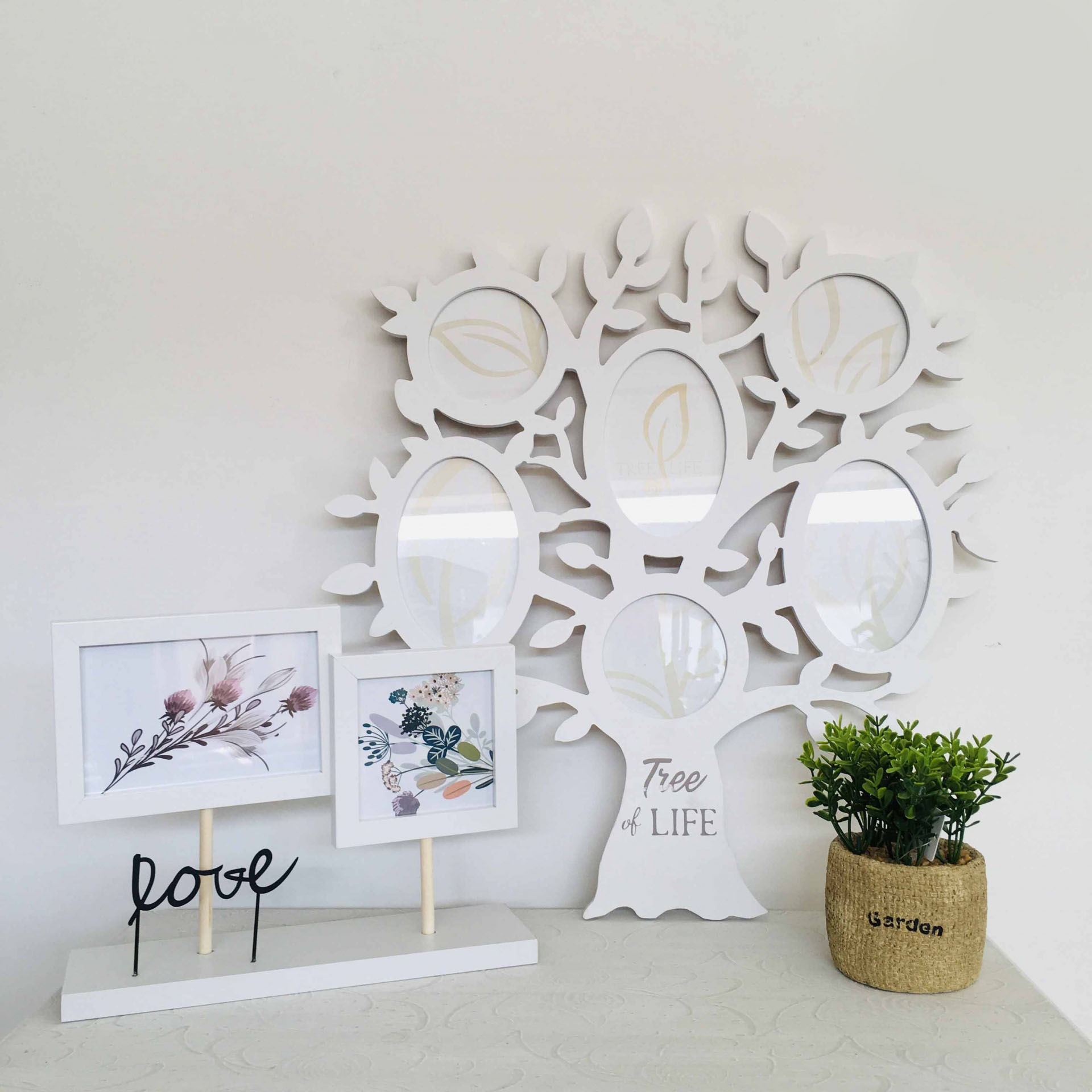 Conjunto decorativo com moldura de árvore branca, fotos de flores, palavra 'love' e planta em vaso de juta.